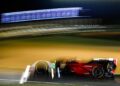 A red Porsche race car speeds on the track at night during Petit Le Mans, creating motion blur with bright light streaks and the Rolex timing banner glowing in the background.