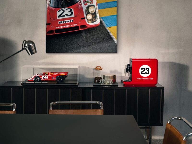 A sideboard with a red SMEG coffee machine, a model race car in a display case, glassware, and a wall photo of a Porsche race car numbered 23.