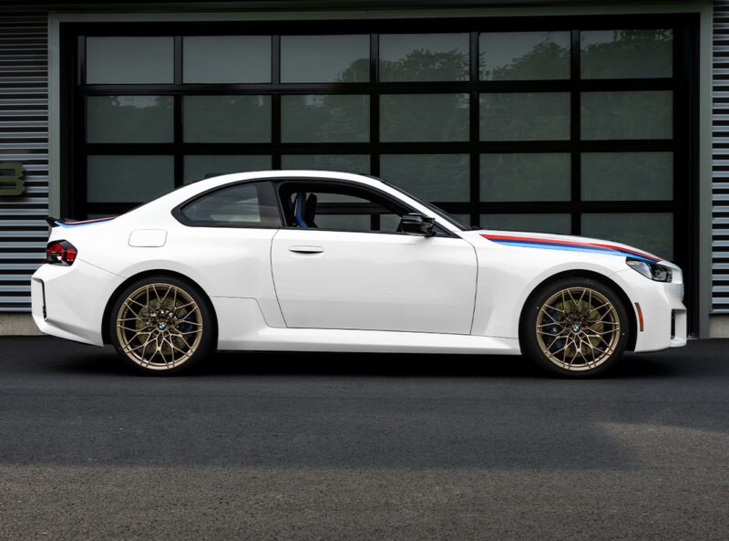 A white two-door sports coupe, inspired by the BMW 2002 Turbo, with gold wheels and blue and red racing stripes is parked in front of a modern glass garage door.