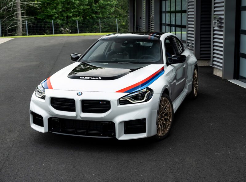 A white 2026 M2 sports car with red and blue stripes is parked on a blacktop next to a modern building and a wooded area, evoking the iconic look of the BMW 2002 Turbo.