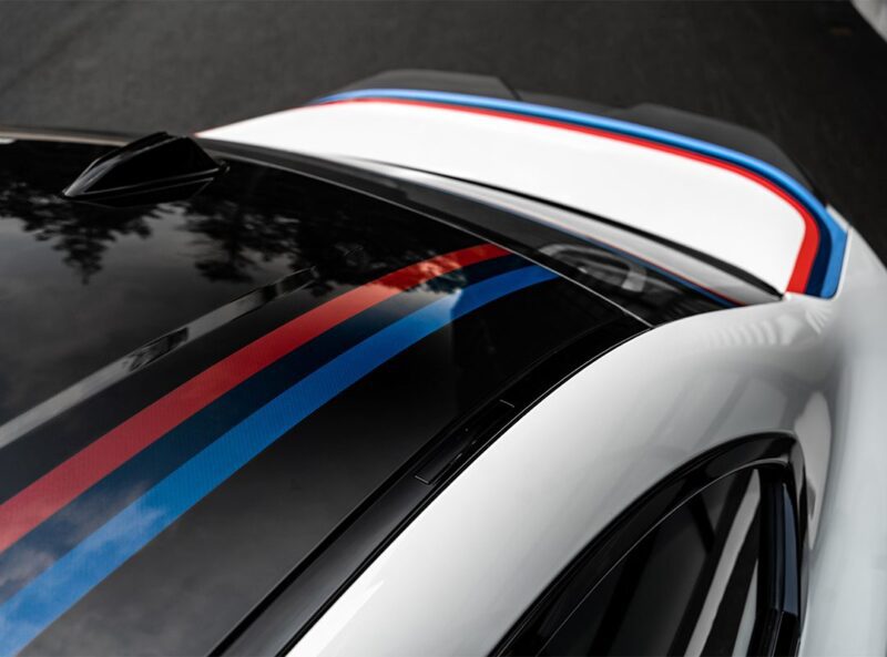 Close-up view of a 2026 M2 Turbo Design Edition’s roof and rear, featuring blue and red racing stripes reminiscent of the classic BMW 2002 Turbo, along with a bold black roof section.