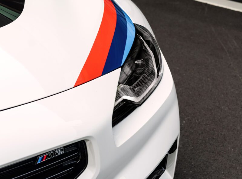 Close-up of a white BMW M2 Turbo Design Edition front end, showcasing the headlight, grille, and signature red, blue, and dark blue racing stripes on the hood.