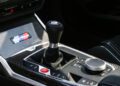 Close-up of a car's interior, featuring the gear shifter, control knob, and a panel with a "turbo" badge on a carbon fiber surface, inspired by the legendary BMW 2002 Turbo and the upcoming 2026 M2 Turbo Design Edition.
