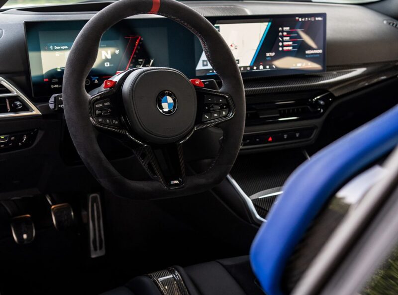 Close-up view of a BMW interior showing the steering wheel, digital dashboard, and part of a blue seat—highlighting details inspired by the Turbo Design Edition for the 2026 M2.
