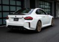A white 2026 M2 Turbo Design Edition with gold wheels is parked in front of a modern building with large glass doors, echoing the spirit of the classic BMW 2002 Turbo.
