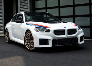 A white BMW 2002 Turbo with blue and red racing stripes and gold wheels is parked on black pavement in front of a modern building with glass doors, showcasing its classic Turbo Design Edition appeal.