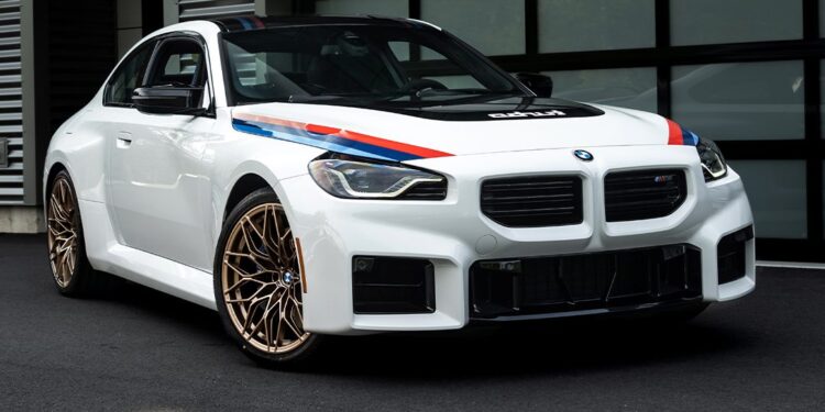 A white BMW 2002 Turbo with blue and red racing stripes and gold wheels is parked on black pavement in front of a modern building with glass doors, showcasing its classic Turbo Design Edition appeal.