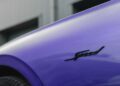 Close-up of a Bentley Continental GTC Speed in striking Violette color, featuring a black "Speed" badge on its side, set against a blurred gray building background.