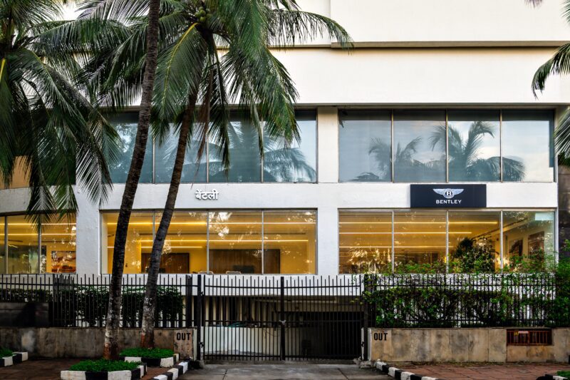 A Bentley showroom with large glass windows, palm trees in front, and a gated entrance labeled “IN” and “OUT,” showcasing the marque’s growing Indian presence.