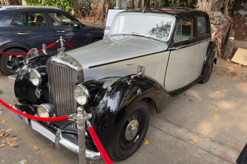 A classic black and silver Bentley vintage car is displayed outdoors, roped off with red barriers, parked beside a modern blue vehicle, highlighting the Indian presence at new showrooms.