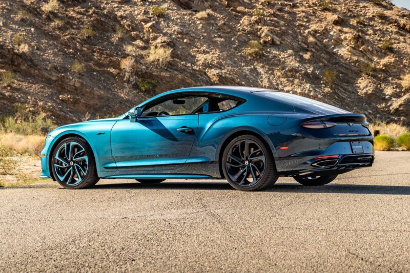 A blue luxury coupe, part of the First Ombré by Mulliner Collection, is parked on a paved road next to rocky desert terrain, viewed from the rear side angle at Bentley Rancho Mirage with three bespoke commissions.