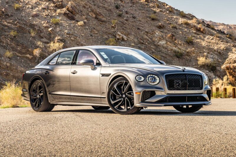 A silver luxury sedan with black wheels is parked on a paved road in front of rocky hills under a clear sky, as the First Ombré by Mulliner Collection arrives at Bentley Rancho Mirage with three bespoke commissions.