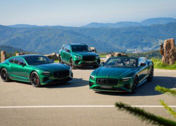Three green luxury Bentley cars—two coupes and one SUV—are parked on a mountain overlook with distant hills and blue sky in the background.