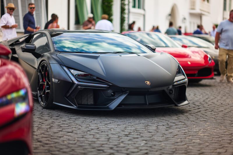A matte black Lamborghini supercar is parked on a cobblestone street among other luxury cars at Alys Beach, drawing attention from several people and car enthusiasts at the Cars of 30A event.