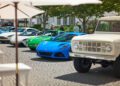 A row of classic and modern supercars, including a blue Lotus sports car, lines a cobblestone street at Cars of 30A, with people and trees in the background.