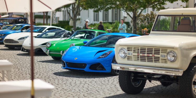 A row of classic and modern supercars, including a blue Lotus sports car, lines a cobblestone street at Cars of 30A, with people and trees in the background.