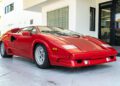 A rare Lamborghini Countach coupe in striking red is parked on a paved surface outside a modern white building with glass doors and windows—an exciting find for those searching for a Countach for sale.