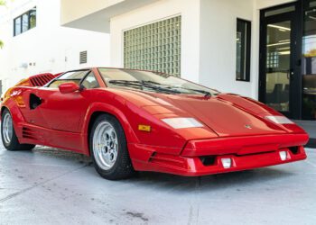 A rare Lamborghini Countach coupe in striking red is parked on a paved surface outside a modern white building with glass doors and windows—an exciting find for those searching for a Countach for sale.