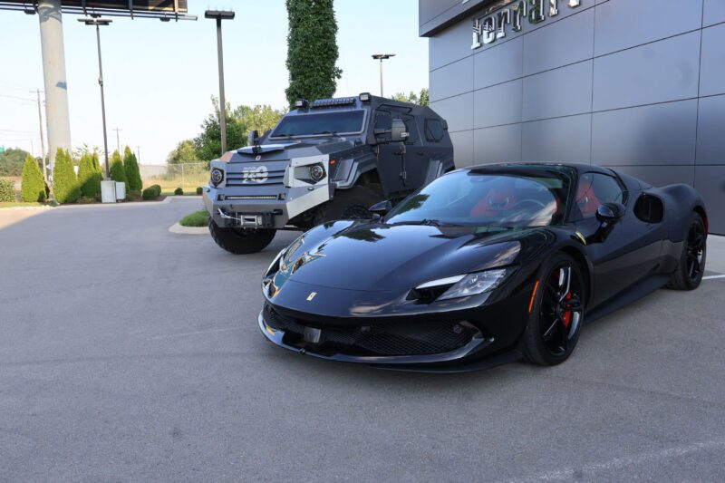 A black Ferrari sports car is parked in front of a matte black armored vehicle outside a modern building with "Ferrari" signage, showcasing Ferrari Nashville’s support for charity fundraising events.