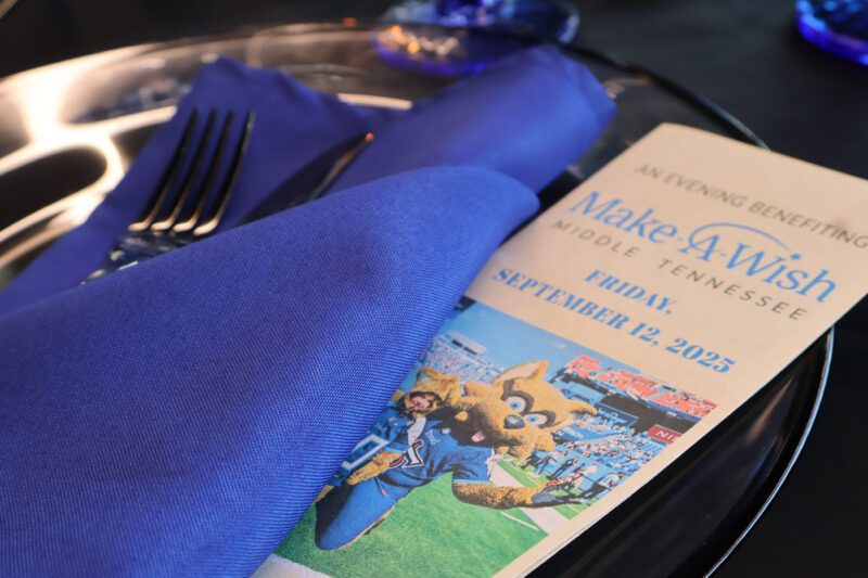 A Make-A-Wish Middle Tennessee fundraising event program for September 12, 2025, rests on a table with a blue napkin, fork, and knife.