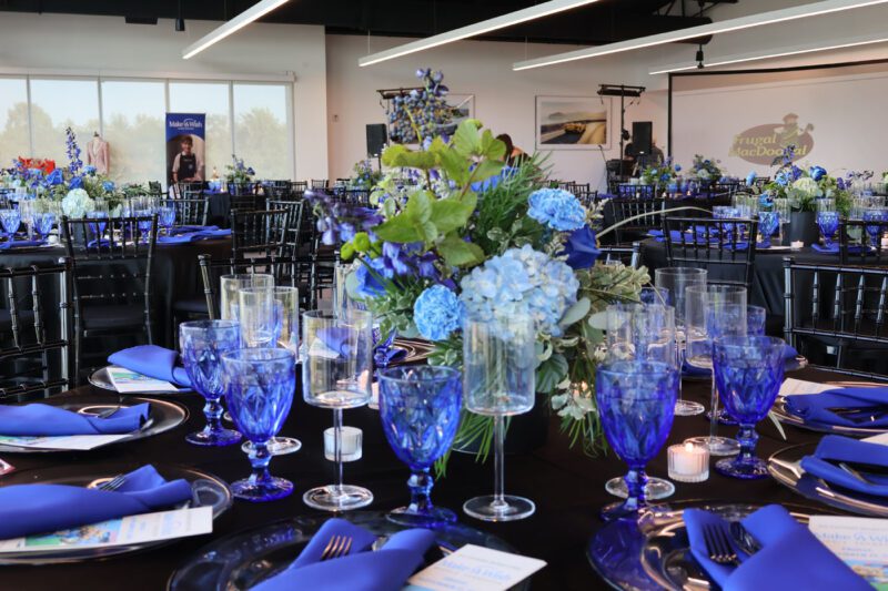 Round tables set for a Make-A-Wish fundraising event feature blue glassware, blue floral centerpieces, navy napkins, and programs at each place. Black chairs and large windows complete the elegant Nashville Ferrari soirée setting.
