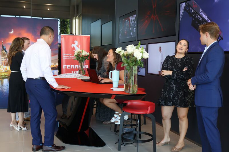 People interact around a red table in a modern office with Ferrari banners, flower vases, and motorsport-themed wall art. One woman uses a laptop while others converse, showcasing the vibrant Ferrari community spirit in Nashville.