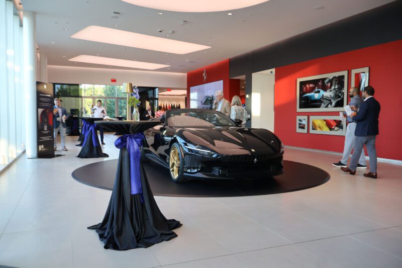 A black Nashville Ferrari luxury sports car is displayed on a circular platform inside a modern showroom during a charity fundraiser, with people standing and conversing nearby.