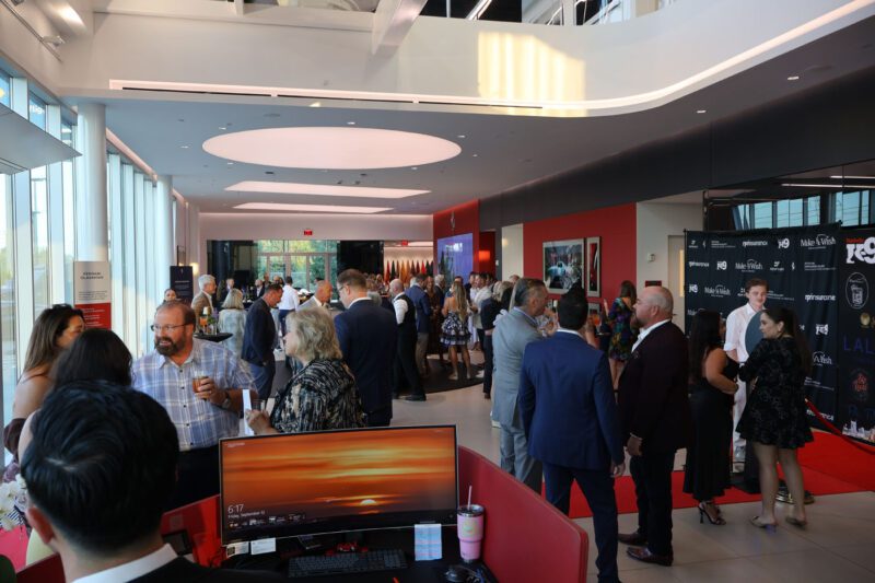 People in formal attire mingle at a Make-A-Wish charity event with large windows, a red accent wall, and a visible red carpet area in the background. A computer monitor sits in the foreground, highlighting Nashville Ferrari’s support.