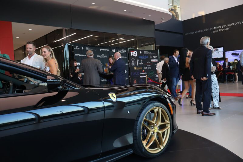People mingle at an upscale charity fundraiser inside a modern showroom, featuring a black Nashville Ferrari in the foreground and a photo wall in the background.