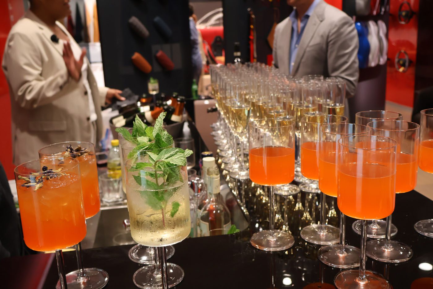 A table with rows of orange cocktails and one clear drink garnished with mint hints at a lively fundraising event; two people in business attire stand behind the table, supporting Make-A-Wish.