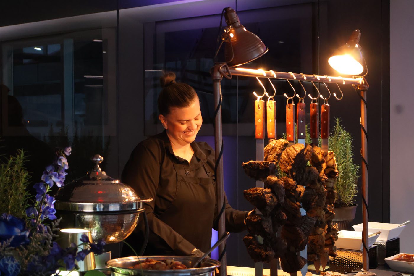 A woman in black attire stands behind a food station with skewered meat and a chafing dish, illuminated by overhead lamps at a Make-A-Wish fundraising event.