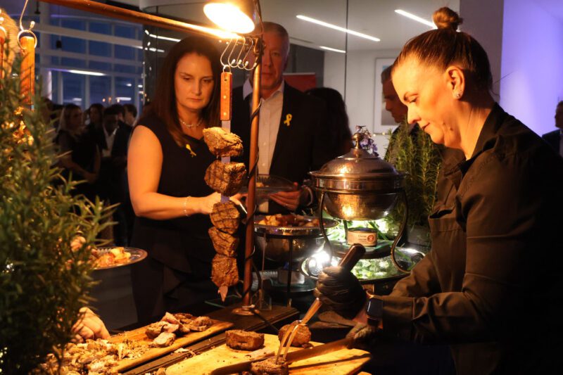 At a charity fundraiser, a woman slices meat from a skewer at a buffet while attendees serve themselves food in the well-lit indoor event setting.