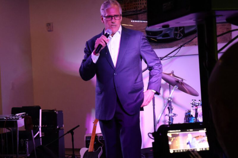 A man in a suit speaks into a microphone on stage at a charity fundraiser, with musical instruments, including a drum set and guitar, visible in the background.