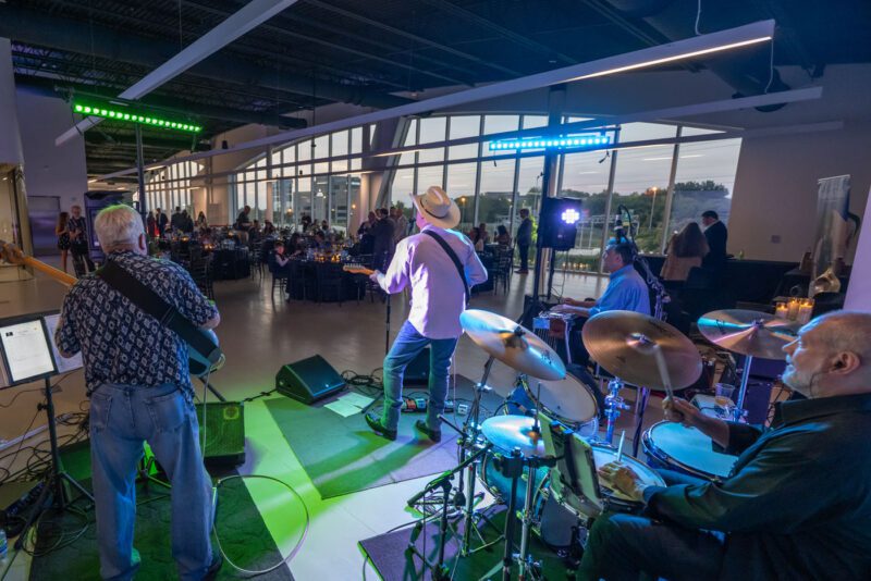 A band performs on stage inside a modern venue with large windows during a Make-A-Wish charity event, while guests are seated at tables in the background.