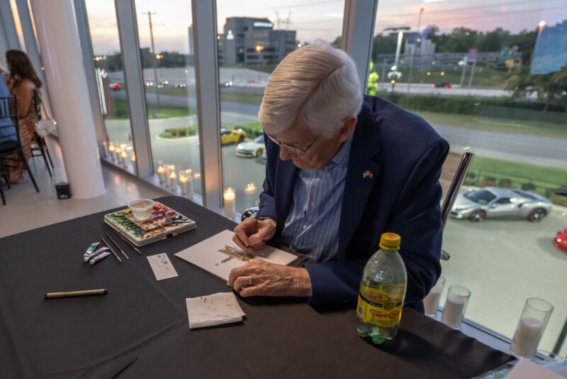 An older man in a suit jacket sits at a table by large windows, drawing on paper with markers to support Make-A-Wish fundraising; a bottle of Topo Chico and art supplies are nearby.