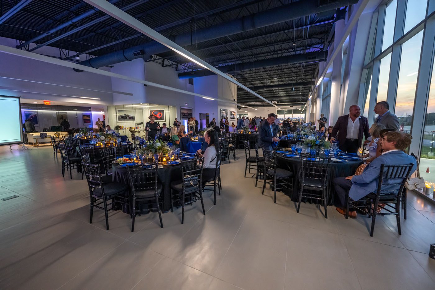 People are seated at round tables set for a charity event in a modern, spacious venue with large windows and bright lighting, supporting Make-A-Wish.