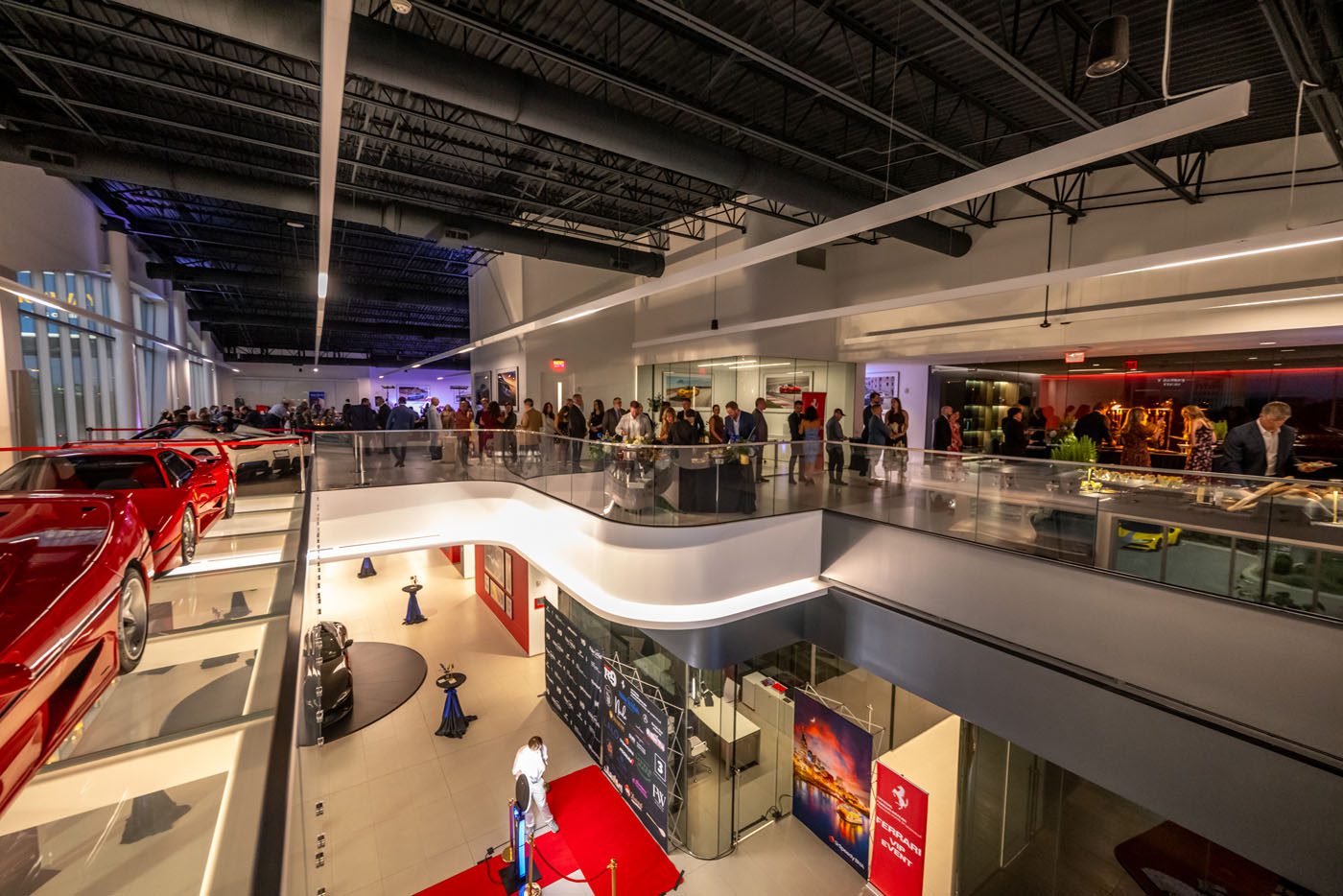 A large crowd gathers inside a modern showroom for a Nashville Ferrari charity fundraiser, with red sports cars on display and a red carpet area on the lower level.