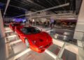 A red sports car is displayed on a glass floor in a modern Nashville showroom, with people from the Ferrari community gathered in the background for a Make-A-Wish event.