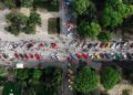Aerial view of parked colorful cars lined up along paths in a park during the Modena Cento Ore 25th Anniversary, surrounded by trees and people walking around the area.