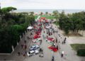 Aerial view of a car show celebrating the 25th Anniversary of Modena Cento Ore, with various sports cars on display, surrounded by people, trees, and a seaside in the background. Event dates: Oct. 5-10.