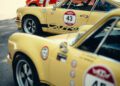 Several vintage yellow Porsche race cars with numbered decals are parked in a row at an outdoor car show, celebrating the 25th Anniversary of the Modena Cento Ore, held from Oct. 5-10, with people and trees in the background.