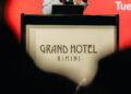 A man speaks into a microphone at a podium labeled "Grand Hotel Rimini," with a red background behind him during the Modena Cento Ore 25th Anniversary, Oct. 5-10.