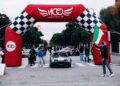 Classic cars drive under a red and black checkered race arch at the Modena Cento Ore 25th Anniversary event, as a person holding an Italian flag directs them outdoors.