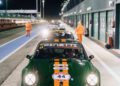 A green race car with orange stripes and the number 44 is parked in a pit lane at night during the Modena Cento Ore, celebrating its 25th Anniversary, with other cars and track personnel in the background.
