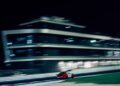 A red sports car races on a track at night with a blurred modern building in the background, celebrating the 25th Anniversary of Modena Cento Ore, held from Oct. 5-10.
