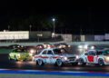 A group of vintage race cars compete on a lit racetrack at night during the 25th Anniversary Modena Cento Ore, with several cars closely navigating a curve in the foreground.