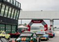Several vintage race cars line up on the track near pit garages, waiting at a starting archway with checkered flags during the Modena Cento Ore’s 25th Anniversary in October 2024.