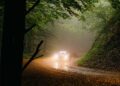 A car with headlights on drives along a winding, leaf-covered road through a forest on a foggy day, evoking the spirit of the Modena Cento Ore 25th Anniversary, held from Oct. 5-10.