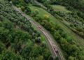 Aerial view of a winding road through a green forested area, with several sports cars—part of the Modena Cento Ore 25th Anniversary event, Oct. 5-10—driving along the curve.