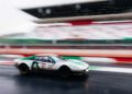 A white race car with green stripes and the number 28 speeds along a wet track at the Modena Cento Ore, celebrating its 25th Anniversary, with blurred grandstands in the background.